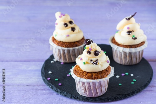 Carrot cupcakes with ghost cream decor and sprinkles on a slate board on a purple wooden background. Happy Halloween concept.