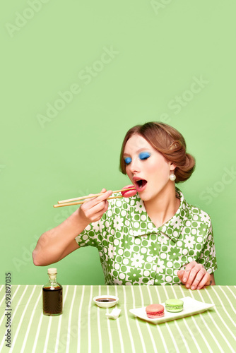 Pretty  stylish  young girl eating sweet macarons with chopsticks against green studio background. Soy sauce taste. Food pop art photography. Concept of retro style  creative vision  imagination.