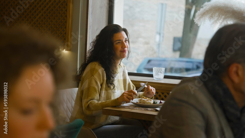 Young woman sits at the table in modern cafe, eats delicious food, works on freelance in laptop. Female freelancer has dinner in restaurant, talks to waiter. Concept of remote work and public catering