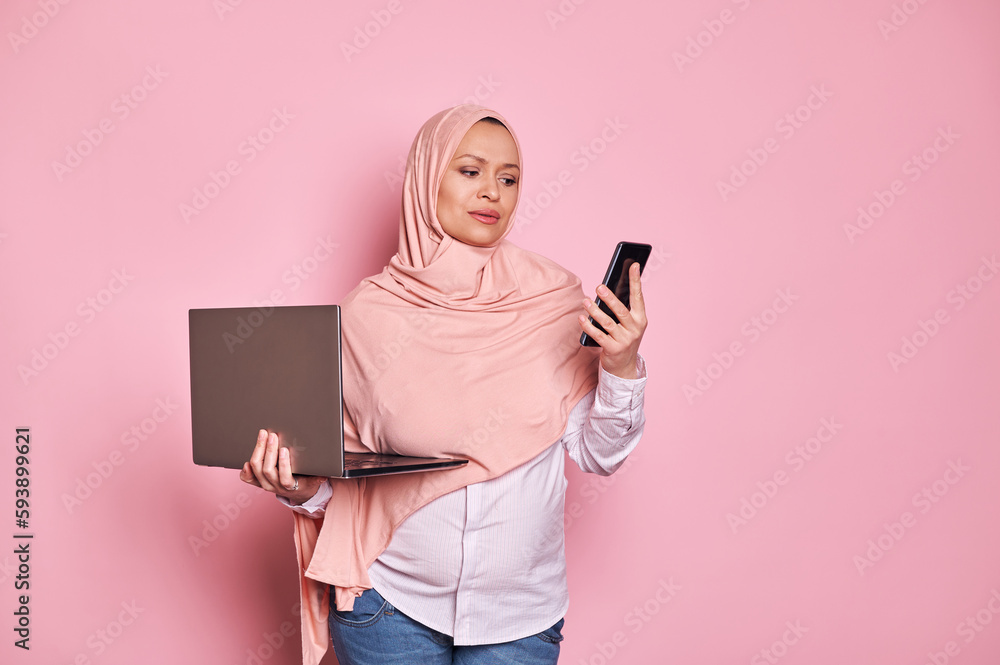 Beautiful Arab Muslim pregnant business woman, wearing pink hijab, holding laptop computer and smart mobile phone, looking perplexed and doubtful, isolated on pink background. Pregnancy. Career. Job Beautiful Arab Muslim pregnant business woman, wearing pink hijab, holding laptop computer and smart mobile phone, looking perplexed and doubtful, isolated on pink background. Pregnancy. Career. Job