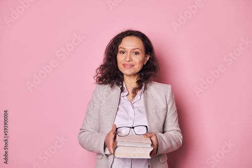 Fotografie Attractive multi-ethnic middle aged woman, school teacher librarian smiling at camera, holding book over isolated pink background