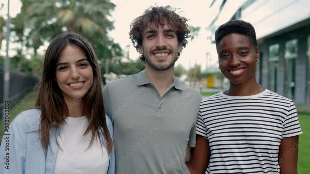 Three multiracial young friends smiling at camera outside. Joyful millennial people standing together hugging each other on summer at city street. Diversity and people concept Three multiracial young friends smiling at camera outside. Joyful millennial people standing together hugging each other on summer at city street. Diversity and people concept