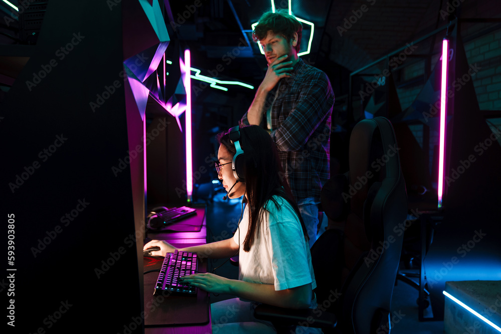 Friend of asian girl watching her playing video game in cybersport club