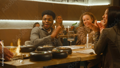 The chef prepares meal in flambe style in front of people in modern gastro cafe. Cheerful multi ethnic friends spend weekend evening, hangout on birthday party in restaurant. Concept of public eating.