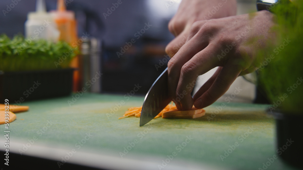 The cook cuts sweet potato on cutting board. Hands of professional chef close-up. Greenery and other ingredients on kitchen desk. Process of cooking gourmet dish. Public catering. Vegetarian dish. The cook cuts sweet potato on cutting board. Hands of professional chef close-up. Greenery and other ingredients on kitchen desk. Process of cooking gourmet dish. Public catering. Vegetarian dish.