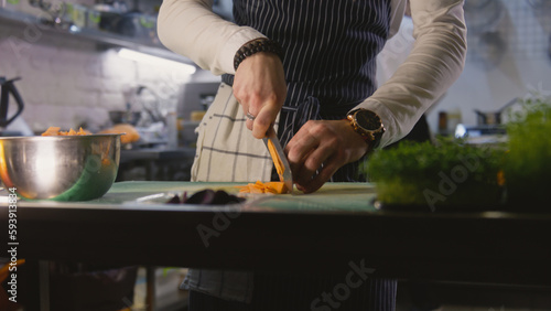 Hands close-up of professional male chef standing by kitchen table and cutting sweet potato. Busy coworker prepares dishes at background. Concept of teamworking in restaurant. Slow motion. Dolly zoom.