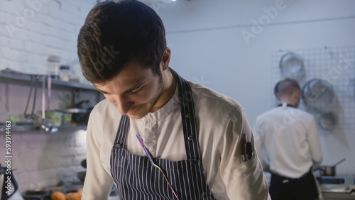Professional chef in apron stands by kitchen table and making food. Colleagues on background preparing ingredients for future dishes. Concept of teamworking in restaurant. Portrait view. Slow motion. © Framestock