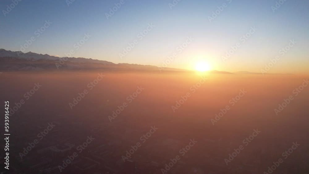 Epic gray smog is visible at sunset over the city. A bird's-eye view from a drone of houses, roads, cars and parks. White clouds and snowy mountains are illuminated by orange rays of the sun. Almaty Epic gray smog is visible at sunset over the city. A bird's-eye view from a drone of houses, roads, cars and parks. White clouds and snowy mountains are illuminated by orange rays of the sun. Almaty