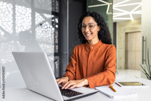 Happy and smiling hispanic businesswoman typing on laptop, office worker with curly hair and glasses happy with achievement results, at work inside office building © Liubomir