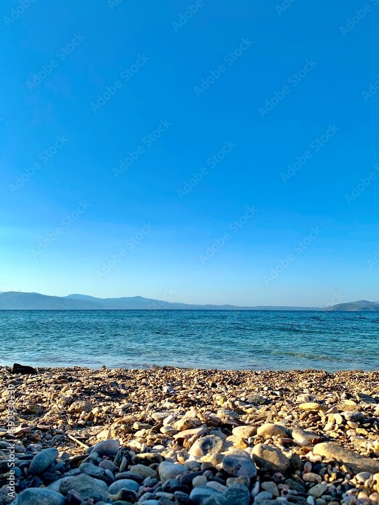 Beach in Greece, mountains in the background. A striking contrast of white walls and green trees, many shades of blue and earth tones under a hazy sky. Walk in nature. Beach in Greece, mountains in the background. A striking contrast of white walls and green trees, many shades of blue and earth tones under a hazy sky. Walk in nature.