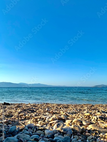 Beach in Greece, mountains in the background. A striking contrast of white walls and green trees, many shades of blue and earth tones under a hazy sky. Walk in nature.  © piece_ov_art