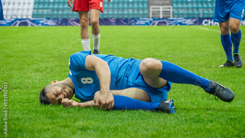 Professional Soccer Football Match Championship: Blue Team Players Attacks, Loses Ball to Foul. Game on an International Tournament. Athlete In Pain Lying on the Grass, Holding a Knee, Struggling