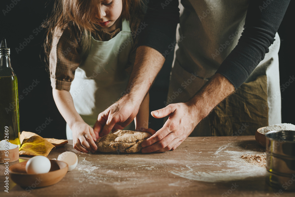 Unrecognizable father and daughter wearing aprons kneading dough on wooden background against dark wall