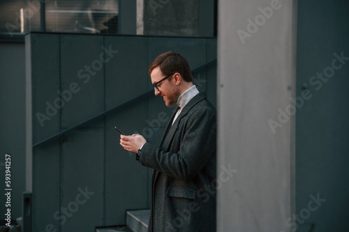 Black colored smartphone in hands. Handsome man in formal clothes is outdoor near the business building © standret