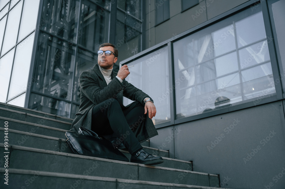 Sitting on the stairs and smoking. Handsome man in formal clothes is outdoor near the business building Sitting on the stairs and smoking. Handsome man in formal clothes is outdoor near the business building