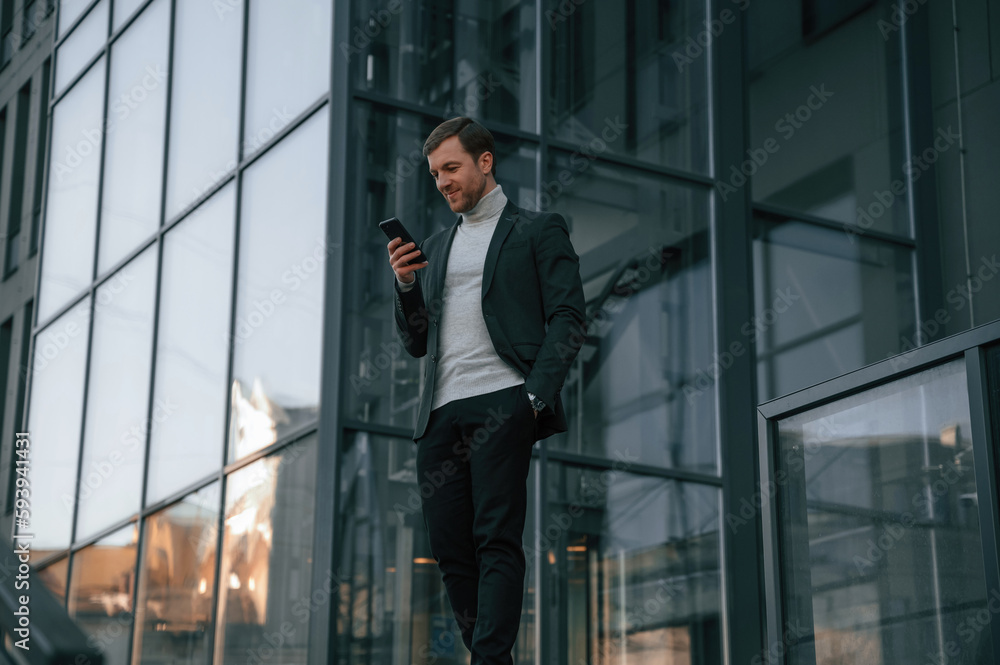 Using social media by smartphone. Handsome man in formal clothes is outdoor near the business building Using social media by smartphone. Handsome man in formal clothes is outdoor near the business building
