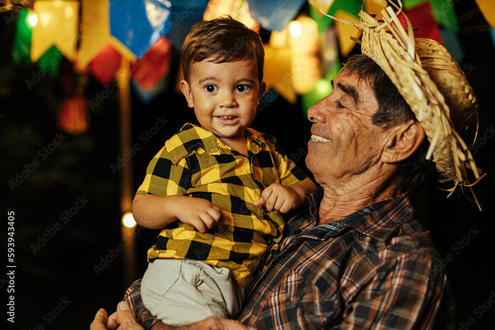 Senior man and his baby grandson celebrating the Brazilian Festa Junina. Portrait of grandfather and his grandson wearing typical clothes and a straw hat during the traditional June festival in Brazil Senior man and his baby grandson celebrating the Brazilian Festa Junina. Portrait of grandfather and his grandson wearing typical clothes and a straw hat during the traditional June festival in Brazil