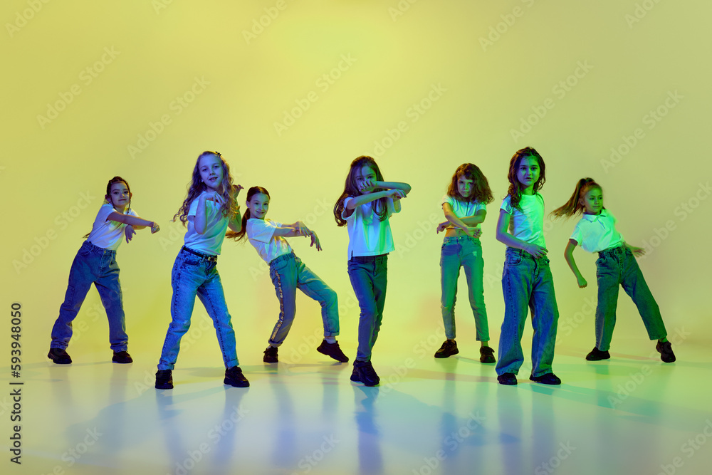 Obraz premium Dynamic image of little girls, children dancing against green studio background in neon light. Casual clothes. Hip-hop style. Concept of childhood, hobby, sportive lifestyle, action and motion