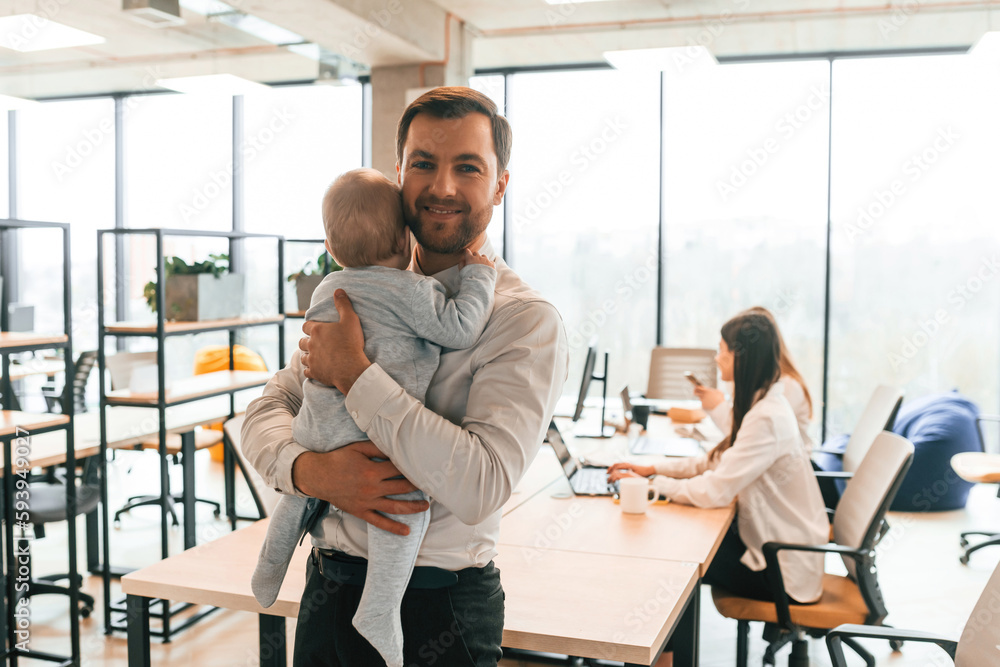 Man with his son. Infant baby is in the office where group of people are working together Man with his son. Infant baby is in the office where group of people are working together