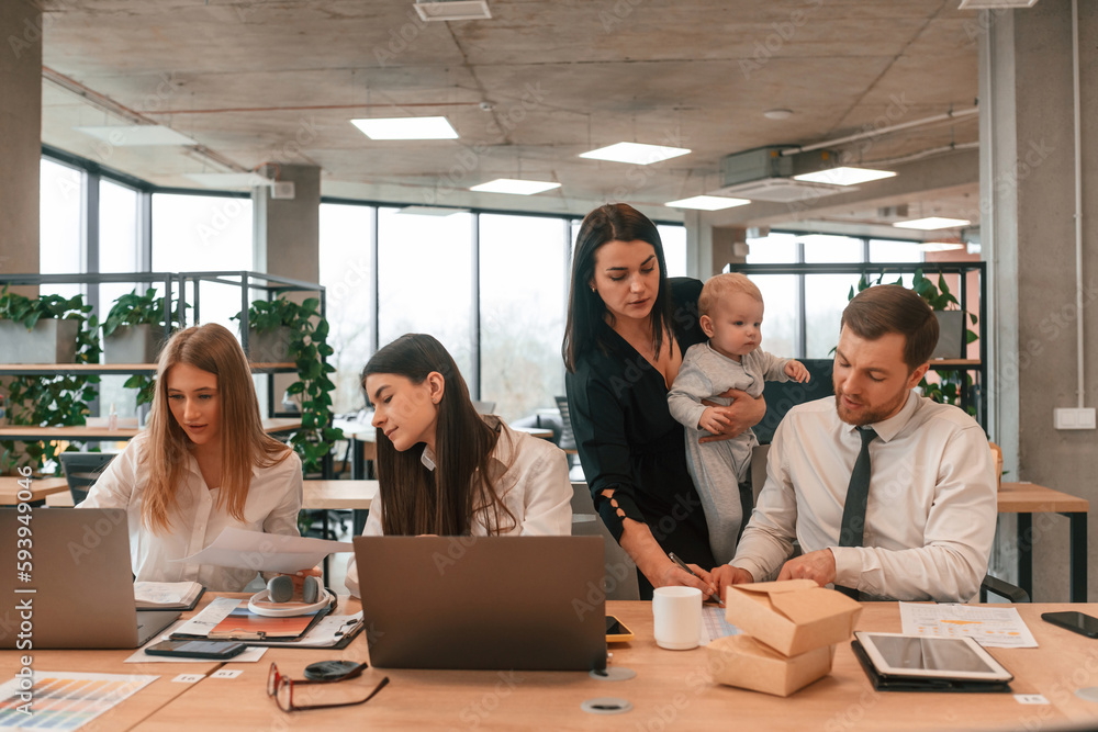 Young mother is holding her son. Infant baby is in the office where group of people are working together Young mother is holding her son. Infant baby is in the office where group of people are working together