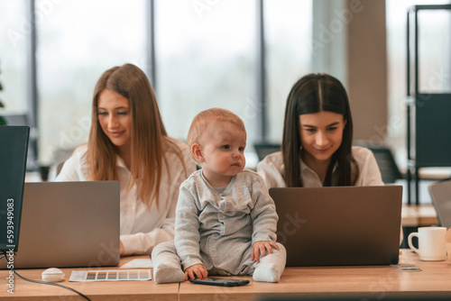 Sitting on the table. Women are using laptops. Infant baby is in the office where group of people are working together