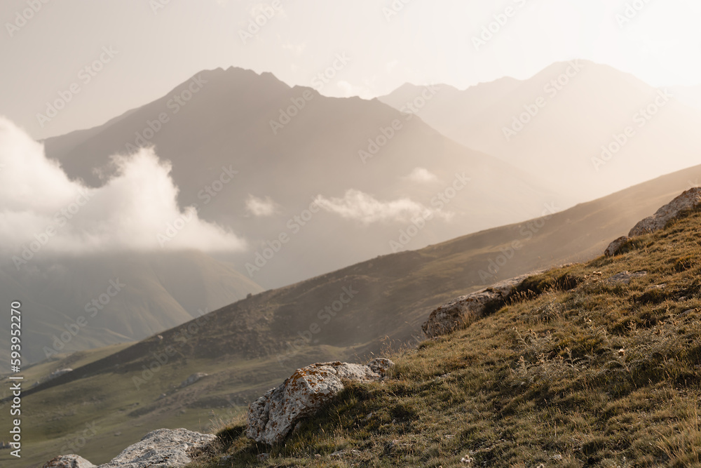 Beautiful mountain landscape - silhouette of high mountain peaks in golden sunbeams of early morning sun, fluffy clouds, white soft mist, slope with dry grass, panorama view on valley in autumn.