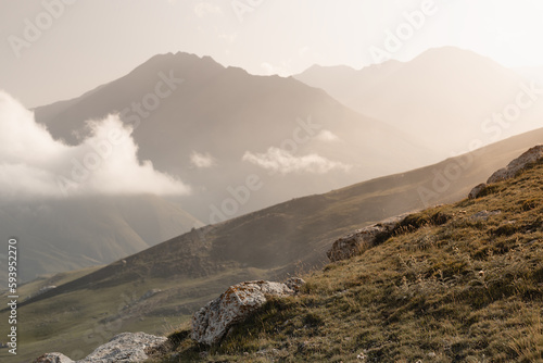 Beautiful mountain landscape - silhouette of high mountain peaks in golden sunbeams of early morning sun, fluffy clouds, white soft mist, slope with dry grass, panorama view on valley in autumn.