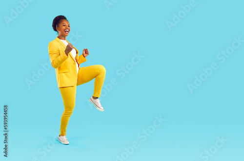 Happy laughing woman dancing cheerfully showing yes gesture clenching her fists isolated on turquoise background, full length. Young stylish african american woman in yellow pants, jacket, sneakers.