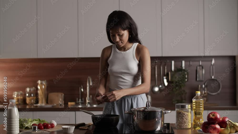 A young black woman cooks herself scrambled eggs for breakfast. A brunette woman standing in the kitchen breaks an egg with a knife and pours it into a frying pan. A young black woman cooks herself scrambled eggs for breakfast. A brunette woman standing in the kitchen breaks an egg with a knife and pours it into a frying pan.