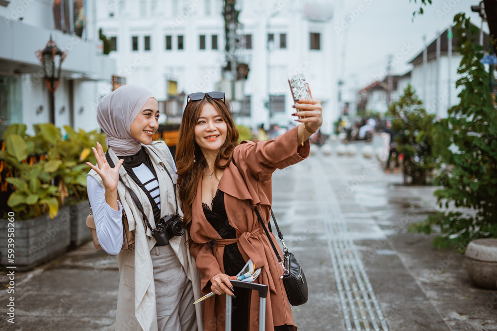 two beautiful travellers taking selfie photo while standing at the sidewalk with the buildings at the background
