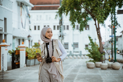 female traveller with hijab smiling while walking through the sidewalk holding the phone and camera
