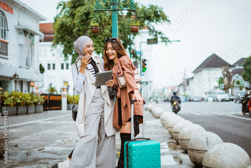 two female travellers laughing while standing together at the sidewalk and looking at the digital tablet © Odua Images