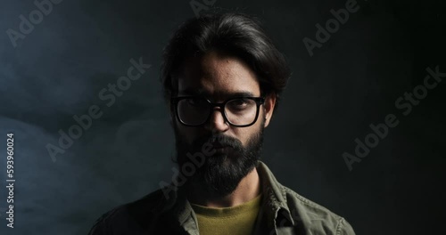 Closeup portrait of serious angry young man with beard and eyeglasses staring at camera against isolated black background photo