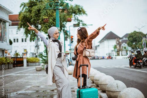 two female travellers pointing to the opposite way while standing together at the sidewalk bring their travel equipment © Odua Images