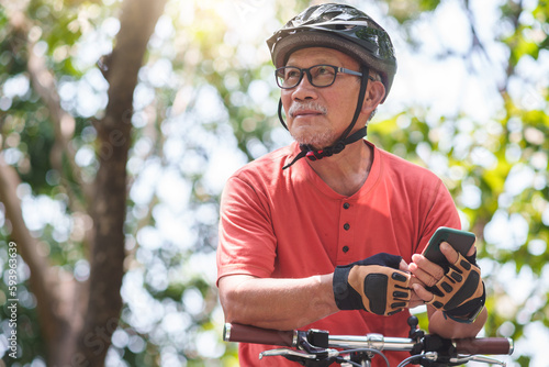 Fotografiet Portrait of Asian Senior Chinese Adult man in helmet holding mobile phone or smartphone while cycling with bicycle at park outdoor