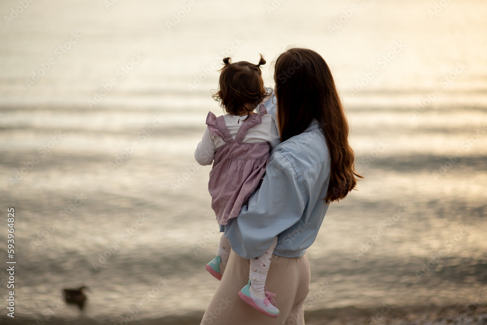 Young brunette mom in blue shirt holding her little toddler girl with two buns in velvet dress at Italian Garda lake beach at sunset Young brunette mom in blue shirt holding her little toddler girl with two buns in velvet dress at Italian Garda lake beach at sunset