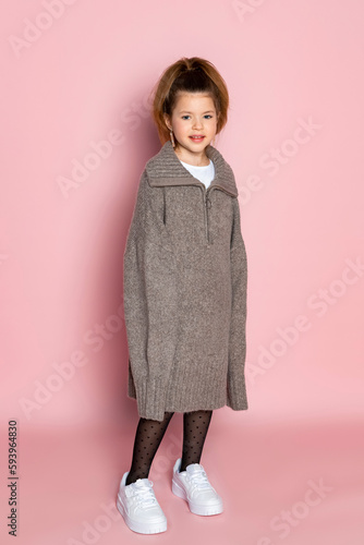 Portrait of a little girl in an oversized brown sweater with a hanging sleeves. Child wearing adult clothes. Isolated on pink background.