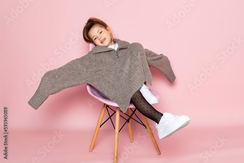 Portrait of a little girl in an oversized brown sweater with a hanging sleeves. Child wearing adult clothes. Isolated on pink background.