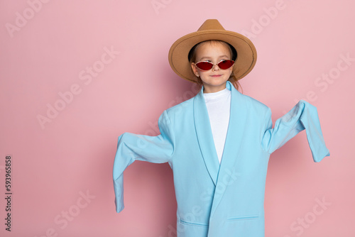Portrait of a little girl in an oversized adult blue blazer, stylish red glasses and a round brown hat. Child wearing adults clothes. Isolated on pink background.