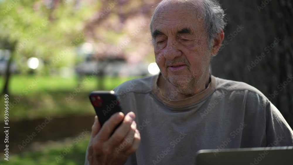 Closeup of elderly old scruffy scruffy man with beard stubble talking to phone having video chat in front of laptop computer outside in a park on a sunny day. Closeup of elderly old scruffy scruffy man with beard stubble talking to phone having video chat in front of laptop computer outside in a park on a sunny day.