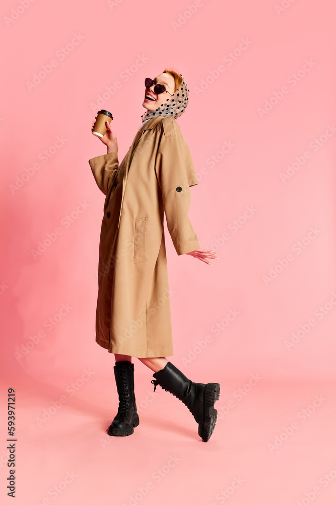 Portrait of charming girl, woman wearing stylish look with headscarf, sunglasses, trench coat holding drink in hand and smiling over pink studio background. Morning coffee