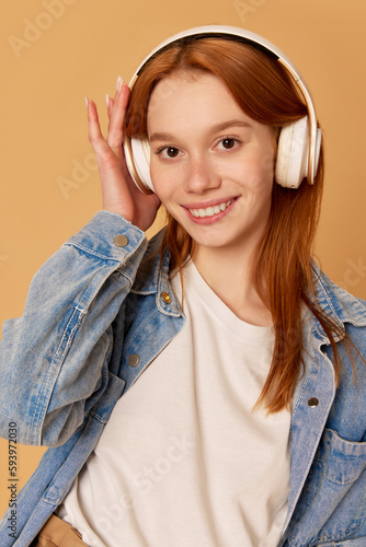 Portrait of young beautiful girl, teenager with red hair wearing headphones and smiling over ginger studio background. Listening to music photo