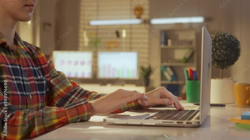 Side view of a young woman counting on a calculator and writing data into a report on a laptop. A woman manager, designer or programmer in a colorful checkered shirt. Close up. Side view of a young woman counting on a calculator and writing data into a report on a laptop. A woman manager, designer or programmer in a colorful checkered shirt. Close up.