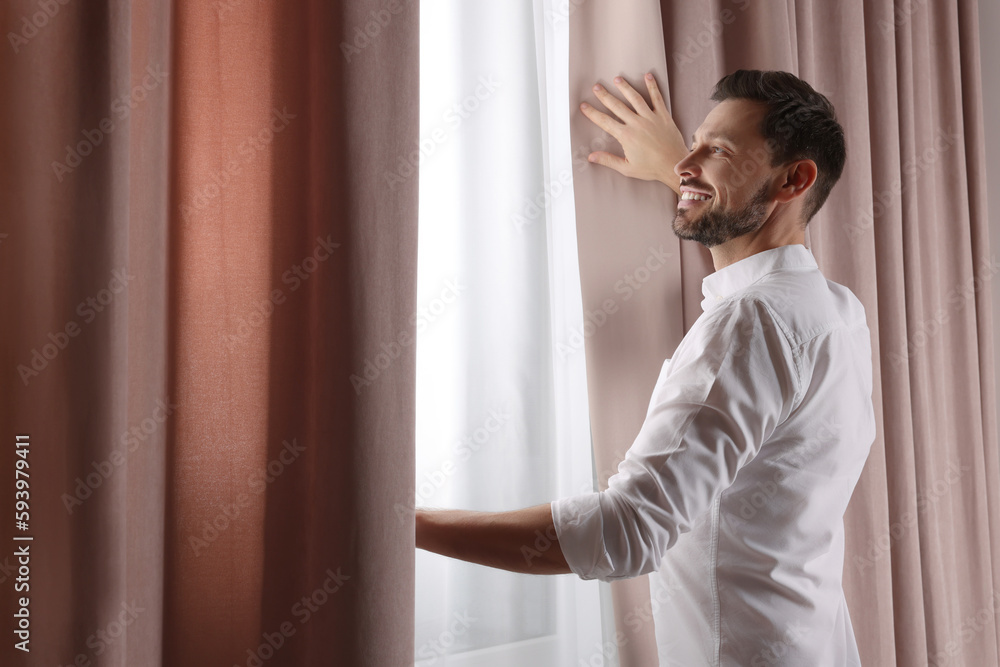 Happy man opening window curtains at home. Space for text