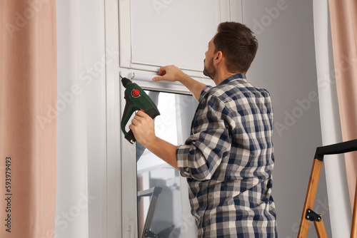 Man with drill installing roller window blind on stepladder indoors