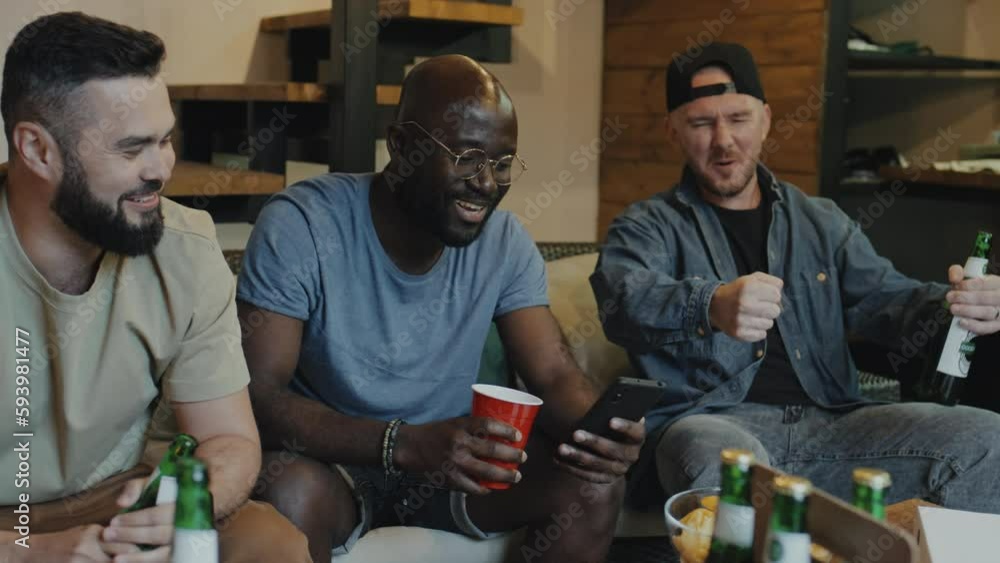 Medium shot of three multiracial friends, two Caucasian men and one black man sitting on sofa with drinks and snacks, watching video on phone, smiling, one imitating car ride Medium shot of three multiracial friends, two Caucasian men and one black man sitting on sofa with drinks and snacks, watching video on phone, smiling, one imitating car ride