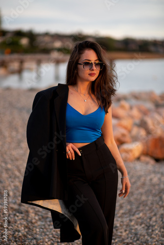 Stylish sexy young brunette girl with sunglasses, black trousers and jacket, blue top posing at sunset light at the lake beach  © Natalia