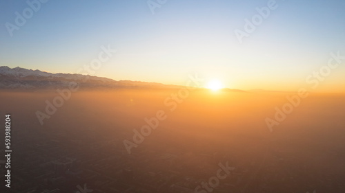 Epic gray smog is visible at sunset over the city. A bird s-eye view from a drone of houses  roads  cars and parks. White clouds and snowy mountains are illuminated by orange rays of the sun. Almaty