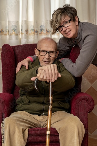 Elder Caucasian man holds a wooden cane while sitting on a couch with her daughter on his side