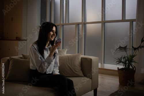 Smiling woman talking on the phone while sitting on the sofa in the living room. A brunette woman sits on a couch in a dimly lit room and records a voice message for her friends.